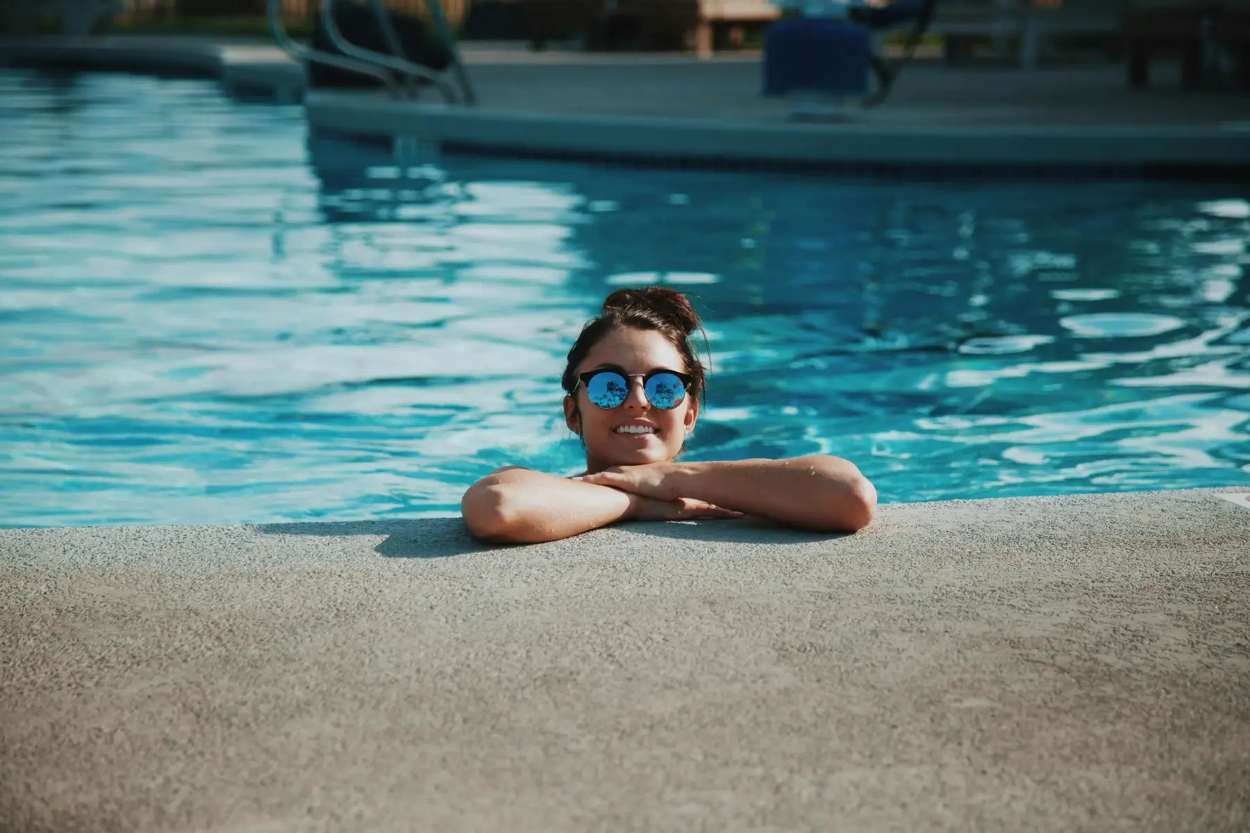 Donna sorridente con occhiali da sole riflettenti che si rilassa a bordo piscina con acqua limpida e trattata, simbolo dell’estate perfetta con i prodotti ChimicaBlu.